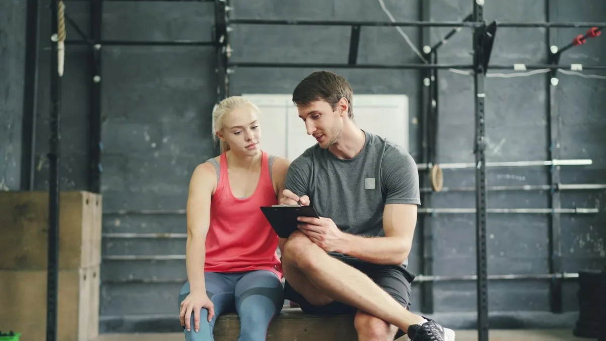 Un istruttore e una cliente seduti su un plyo box in palestra guardano insieme gli appunti su una tavoletta portablocco